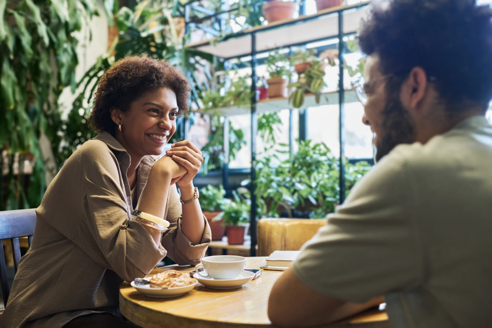 Happy young woman looking at her boyfriend with toothy smile while both sitting in front of each other by table and enjoying date in cafe