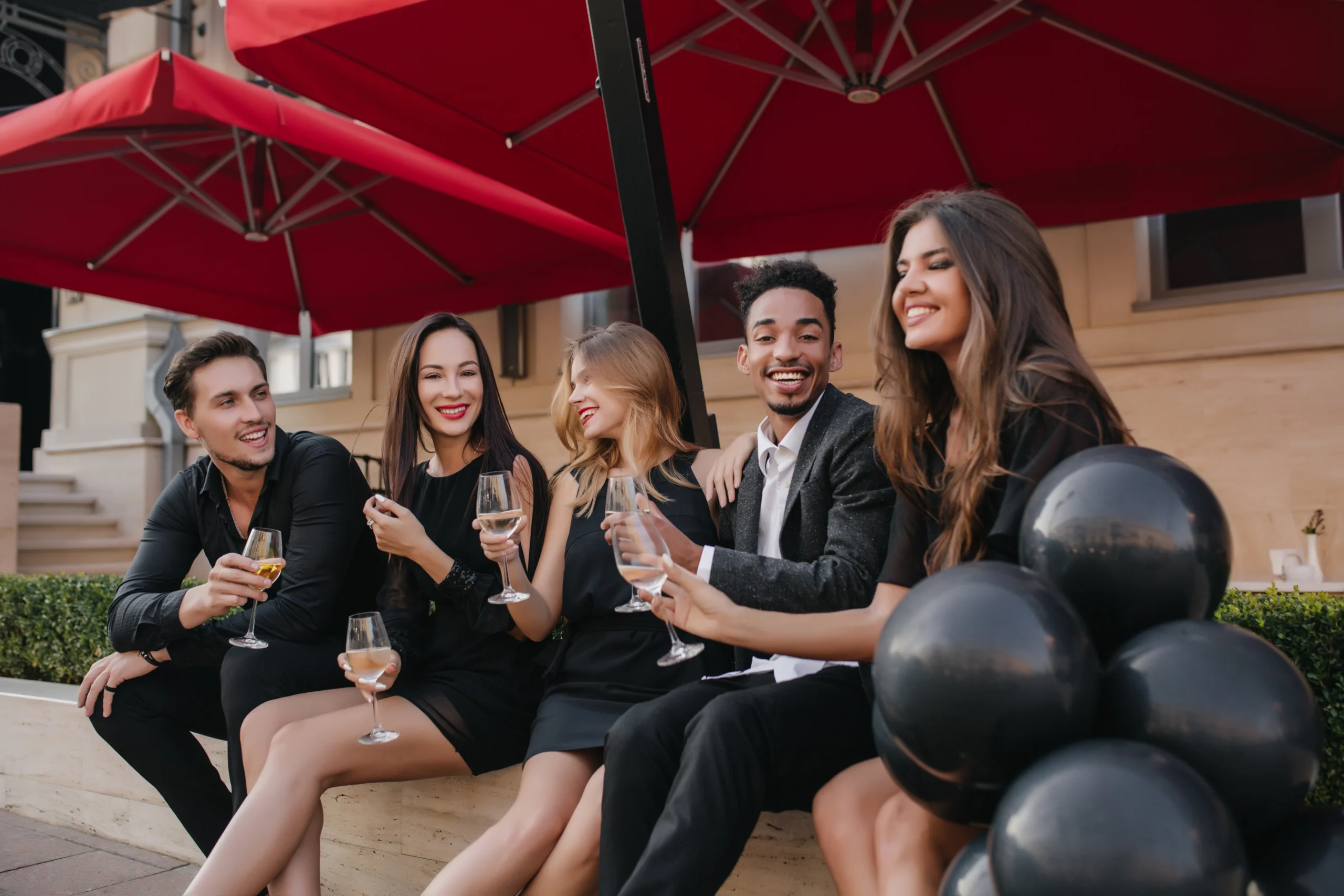 Dreamy girl with light-brown hair sitting in outdoor cafe with black helium balloons. Portrait of handsome african man spending time with european friends in restaurant and drinking wine.