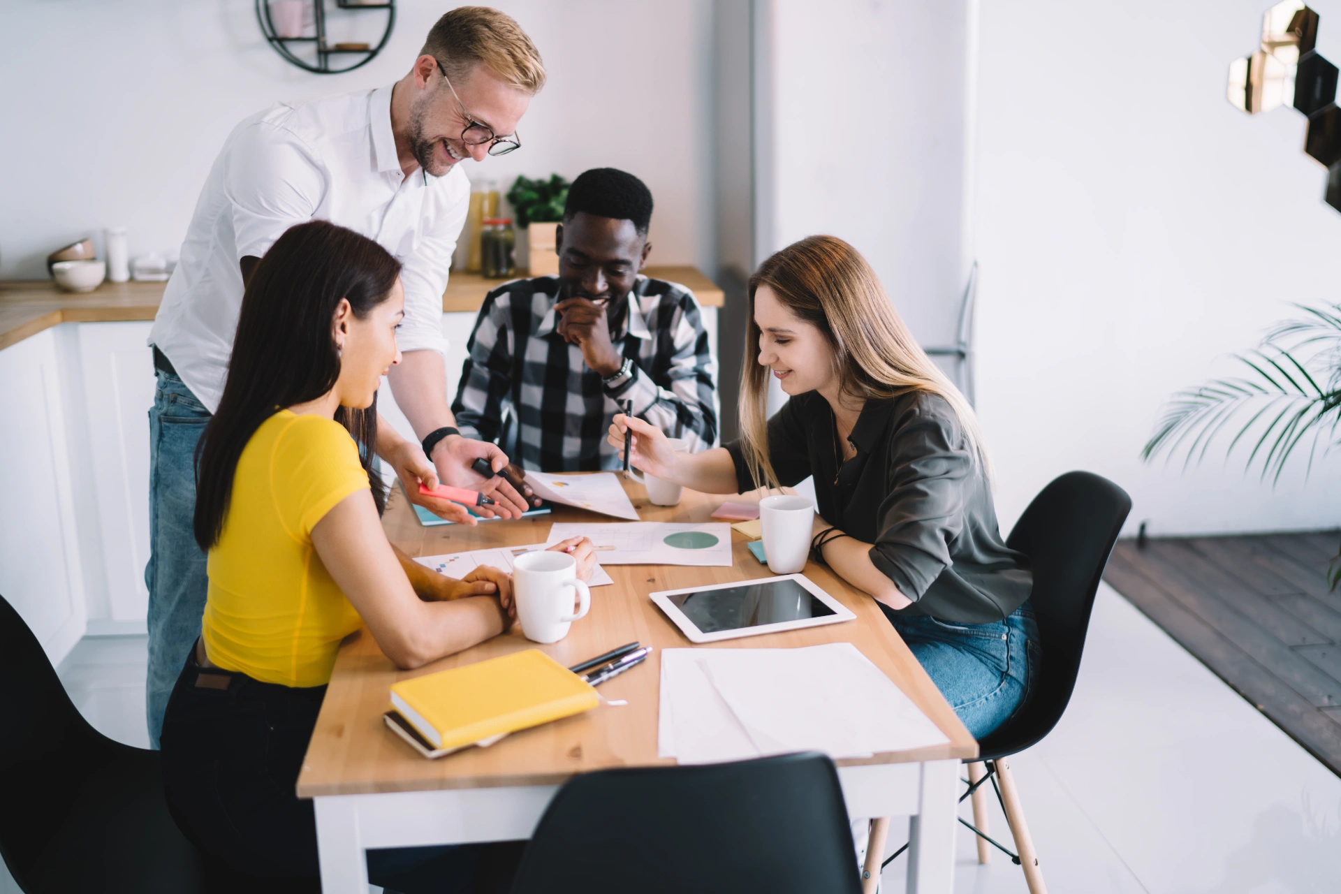 High angle of cheerful multiracial coworkers in casual clothes smiling and working with documents while having meeting at wooden table in kitchen